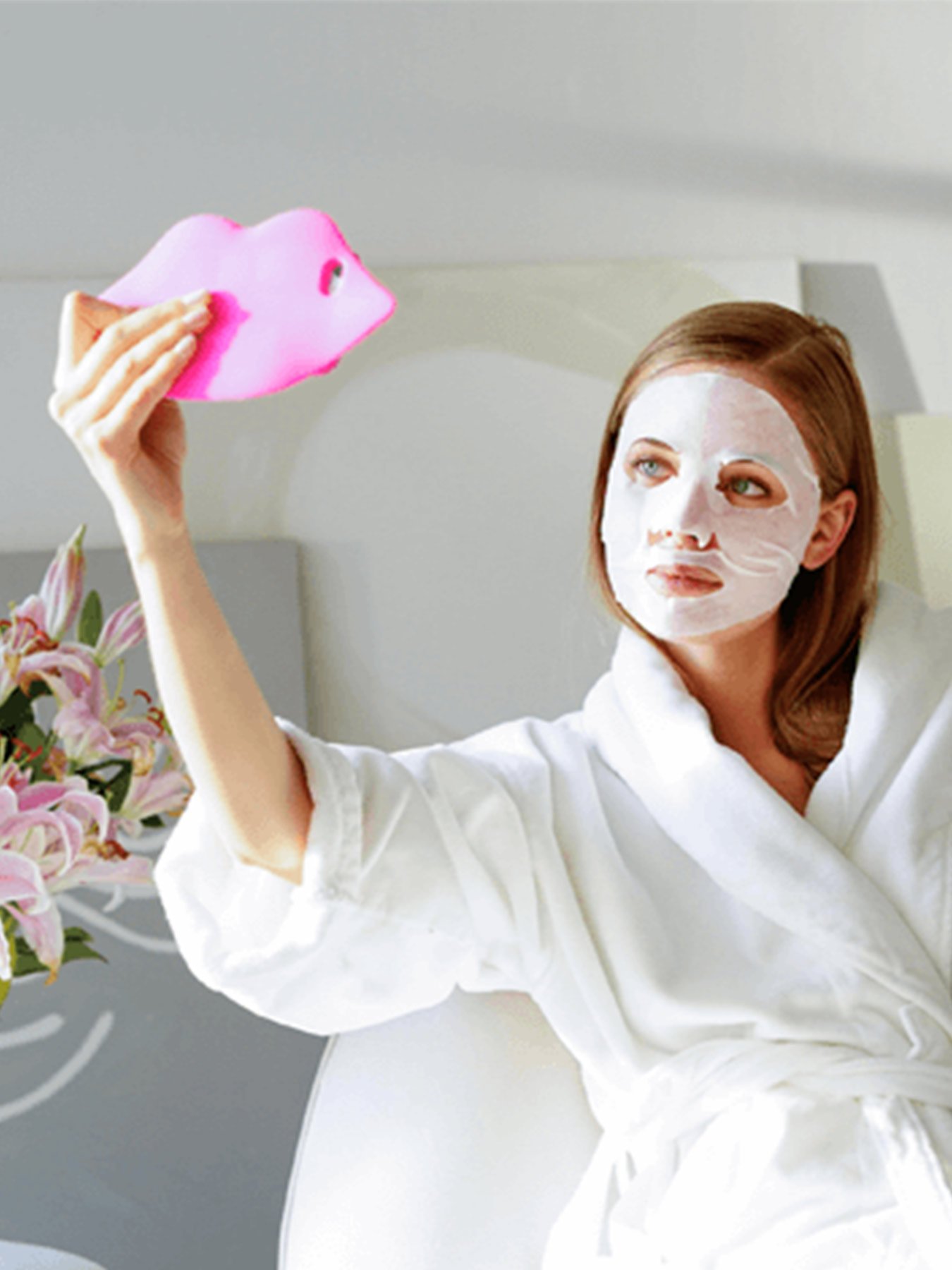 A woman wearing a white bathrobe and The Super Hydrating Sheet Mask - Anti-Fatigue sitting on a white chair holding up a pink lip-shaped mirror in a white and gray room with pink and white flowers on a reflective table.